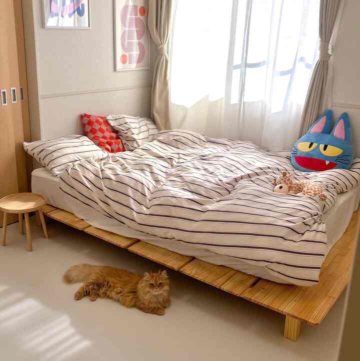 明るい木目調と白を基調とした寝室、ストライプ柄の掛け布団と猫がいる落ち着いた空間