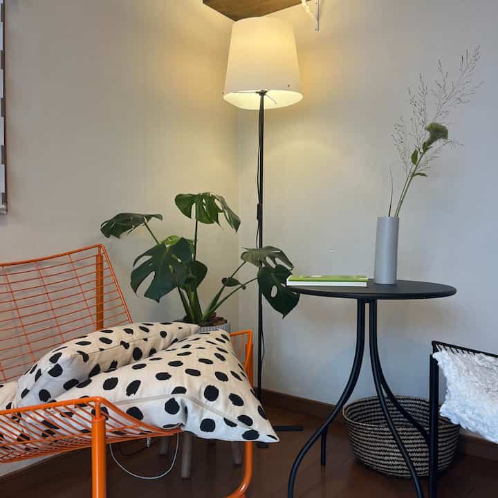 Natural color walls and wood tone floor in a living room corner featuring orange metal armchair with polka-dot cushions, floor lamp, side table, and plant in a simple space