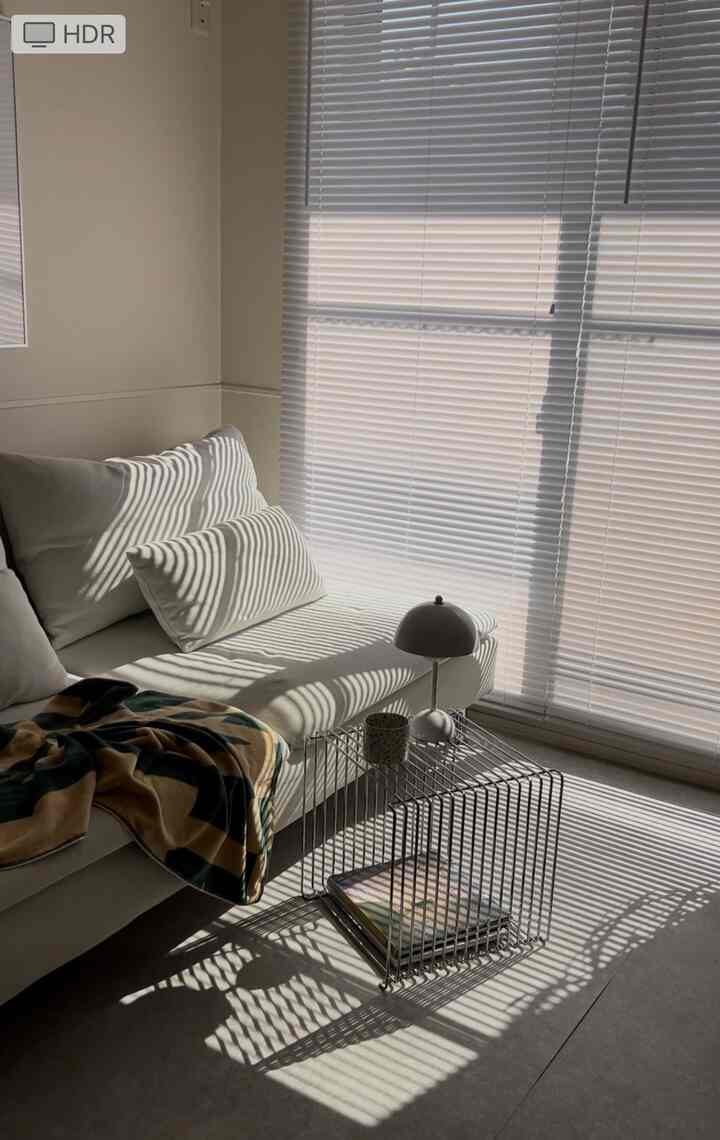 白を基調としたシンプルなリビング、ソファと金属製ブラインドが自然光の影を作り出す落ち着いた空間