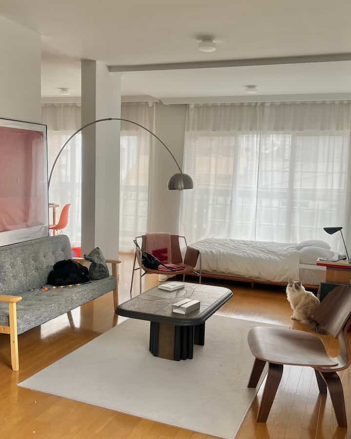 ナチュラルな色調の広々としたワンルーム、グレーのソファと木製のコーヒーテーブルがライトラグに置かれ、窓際にベッドが配置された居心地の良いインテリア