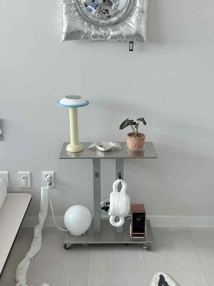 シルバーとホワイトトーンの寝室のナイトテーブル空間、ステンレスのサイドトロリーと照明・植木鉢が配置されたモダンで清潔感のあるインテリア