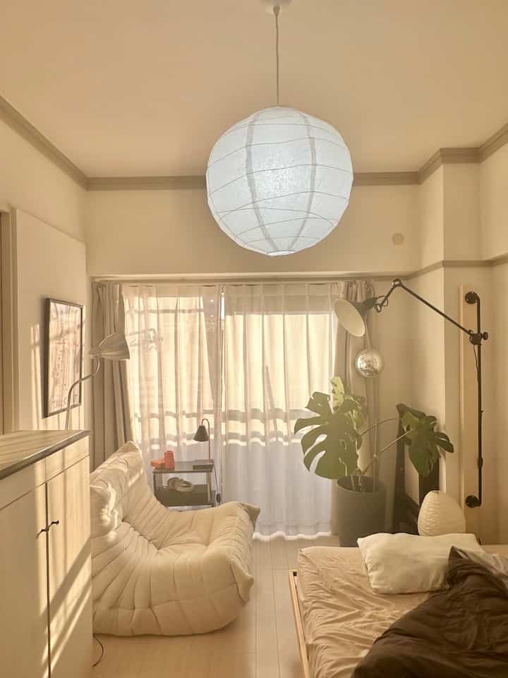 ベージュ調の自然な色合いのベランダ横の一人暮らし向け寝室兼リビング、ソファベッドとペンダントライトが配置された落ち着いたインテリア