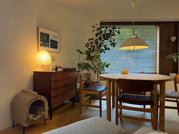 Warm natural-tone living room featuring a wooden round dining table, vintage chest of drawers, cat house, and window blinds