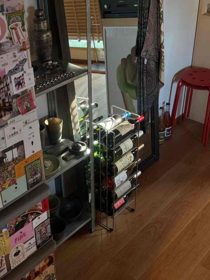 木目調の床に様々なワインボトルが並ぶワインラック、金属製の棚と赤いスツールが配置された一人暮らしのホームバー空間