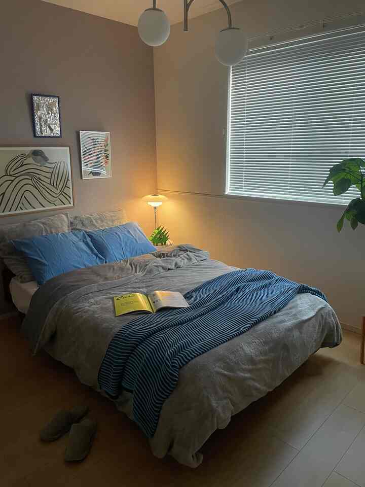 A cozy bedroom with muted beige and gray tones featuring a bed with blue pillows and throw, warm lighting, and minimalistic decor