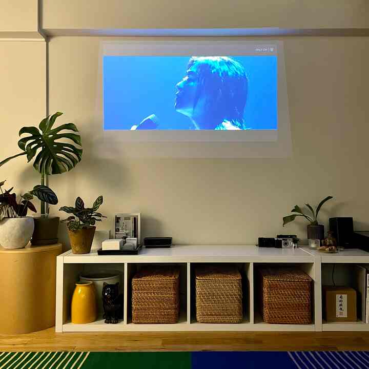 白い壁と棚が配されたミニマルなリビング、ラタンバスケットと植物が調和するナチュラルな雰囲気、中央の壁にプロジェクタースクリーンが映し出されている