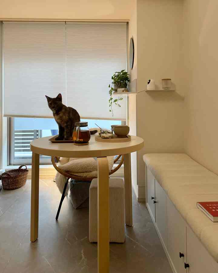 白とナチュラルな色調のリビング、丸い木製テーブルの上に猫が座り、窓には白いブラインドが配置された清潔感のある空間