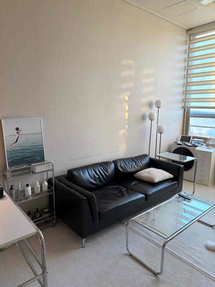 明るいベージュの壁と床に、黒のレザーソファ、透明なガラステーブル、ステンレス製家具が配置されたミッドセンチュリーモダンのワンルーム