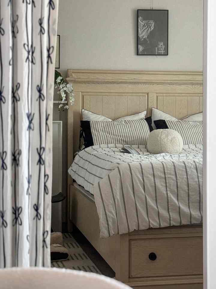Natural-tone bedroom featuring wooden bed frame, striped pillows and duvet, and bow-patterned curtains creating a cozy atmosphere