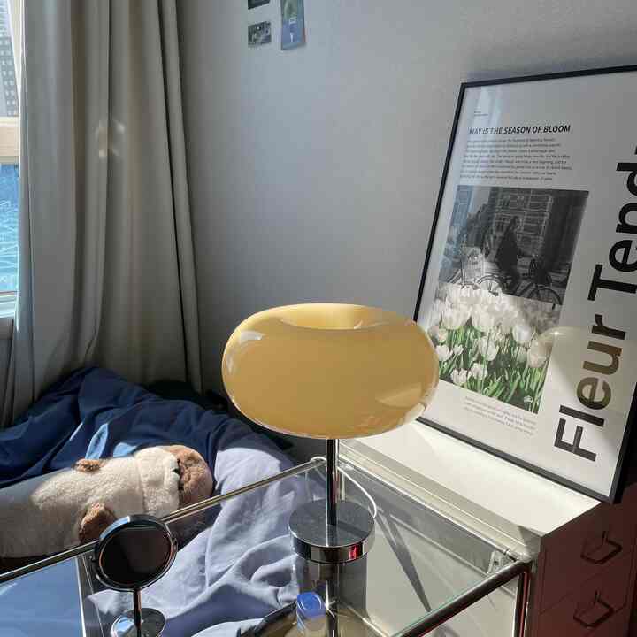 クリームホワイトのカーテンと青い寝具、黄色いドーナツ型のテーブルランプがある居心地の良いワンルームの寝室