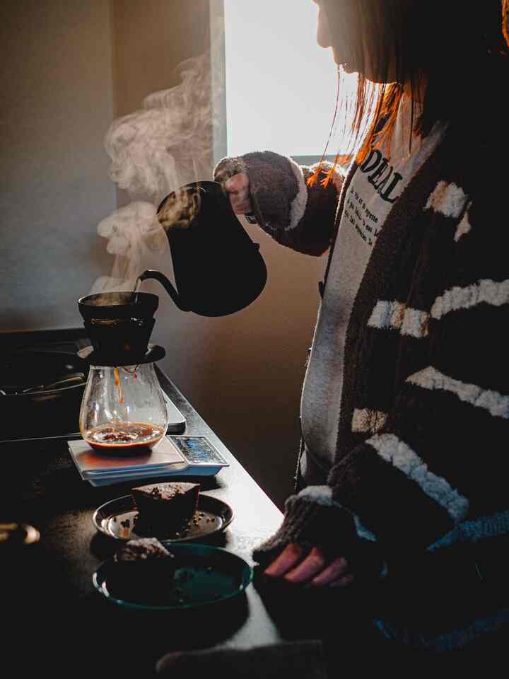 Brown-toned kitchen space featuring a person brewing drip coffee by a window, creating a cozy home cafe atmosphere