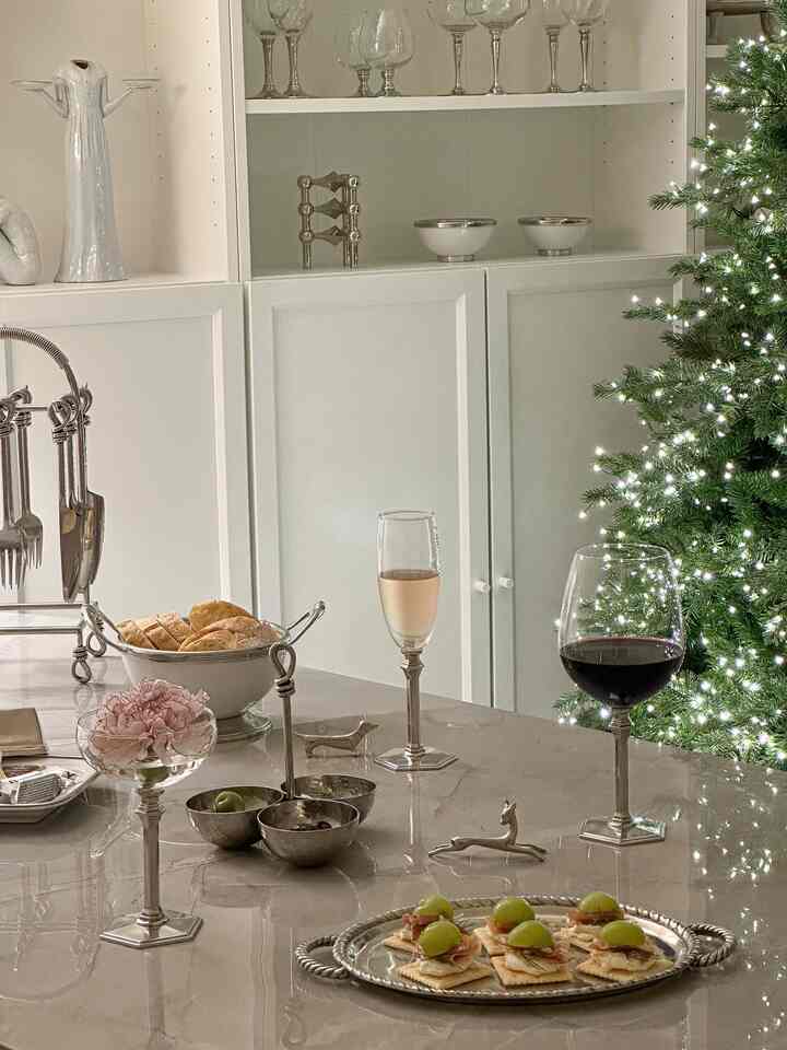 White and silver-toned dining room featuring a Christmas tree, dining table with wine glasses and snacks in an elegant setting