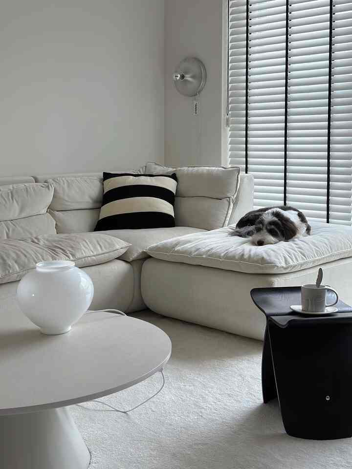 Beige-toned living room featuring a large sectional sofa, vertical blinds, a silver wall sconce, and a white glass vase creating a cozy atmosphere