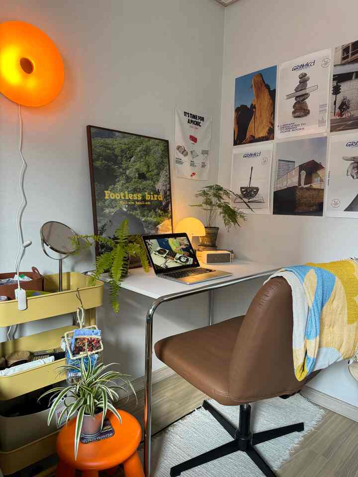 明るいホワイトトーンの壁とテーブルがあるミッドセンチュリーモダンスタイルの書斎、ブラウンの椅子とカラフルなポスター、植物が配置された個性的なデスクインテリア