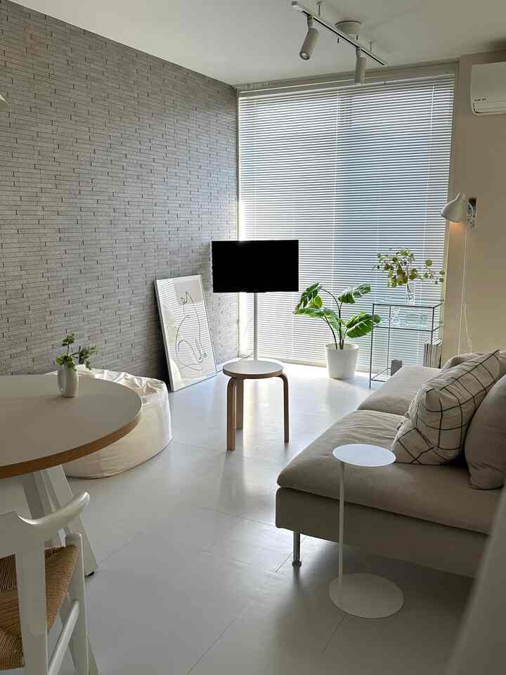 Monotone and white-toned living room featuring beige sofa, round dining table, blinds, and plants in a simple, natural setting