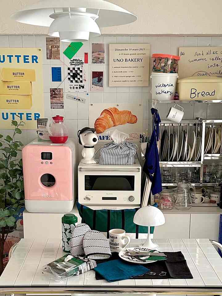 白とベージュのトーンのキッチン空間、中央にオーブンとピンクの家電があり、タイルのダイニングテーブルの上に様々なキッチン用品とテーブルランプが置かれた雰囲気のあるインテリア
