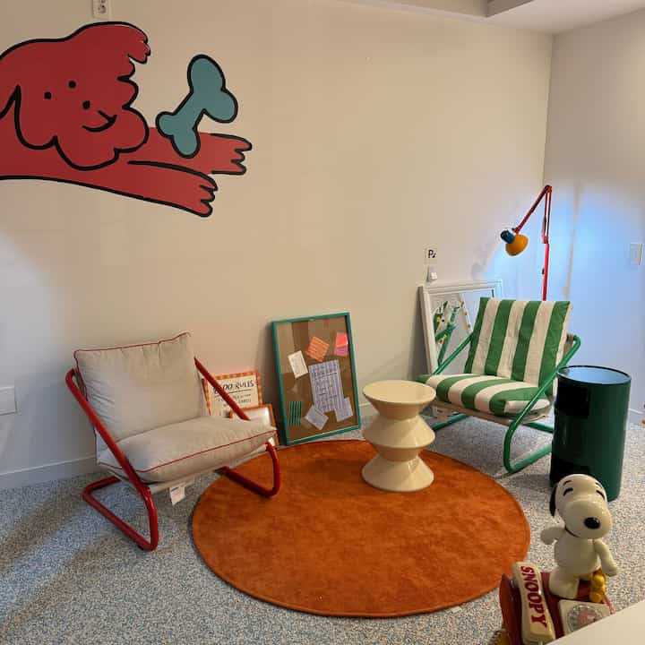 オレンジ色の円形ラグが敷かれたリビングルーム、赤と緑のメタルフレームアームチェアが配置され、壁には犬の絵が描かれた雰囲気のあるインテリア。