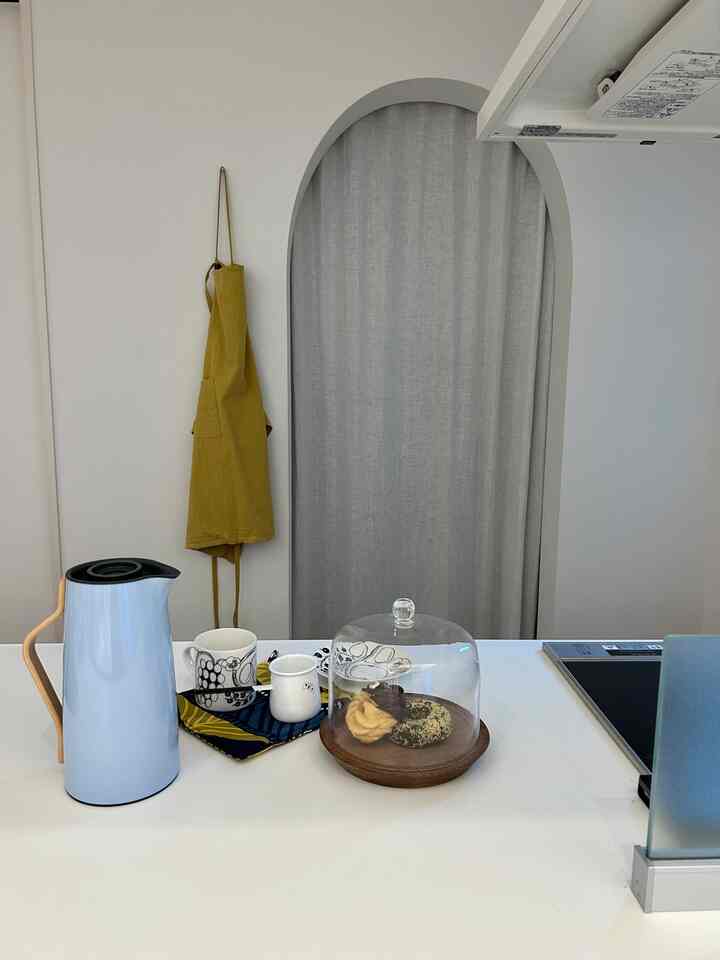 Bright white-toned kitchen space featuring a yellow apron, coffee pot, and cookies under glass dome creating a cozy home cafe vibe