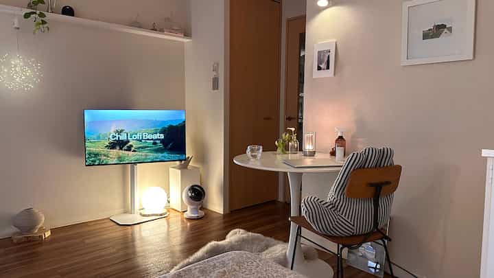 白と木目調が調和したコンパクトなリビング兼ホームオフィス、丸テーブルと快適な椅子が配置され、暖かい照明が雰囲気を演出するインテリア。