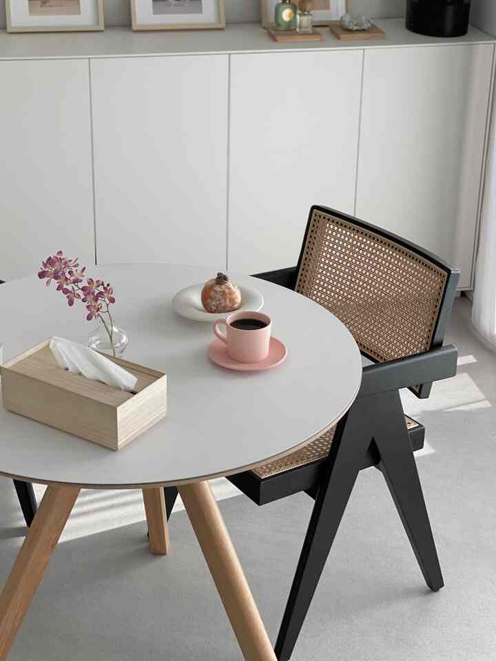 Simple natural dining room with white round table, wood-toned legs, and black rattan chair in a cozy home cafe setting