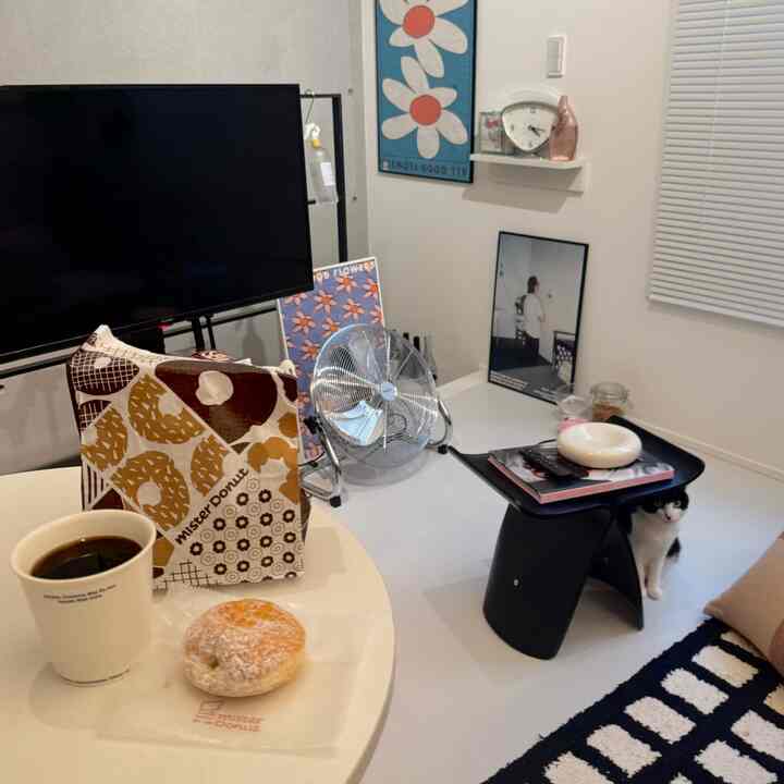 白を基調とした一人暮らしのリビング、黒いスツールの横に猫が座り、丸いテーブルの上にコーヒーとドーナツが置かれた雰囲気のあるおうちカフェ空間