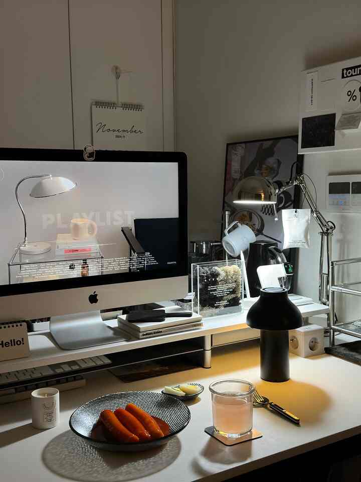 モノトーンの白と黒を基調としたコンパクトなホームオフィス、机の上にデスクランプやiMac、軽食と飲み物が置かれた印象的な空間