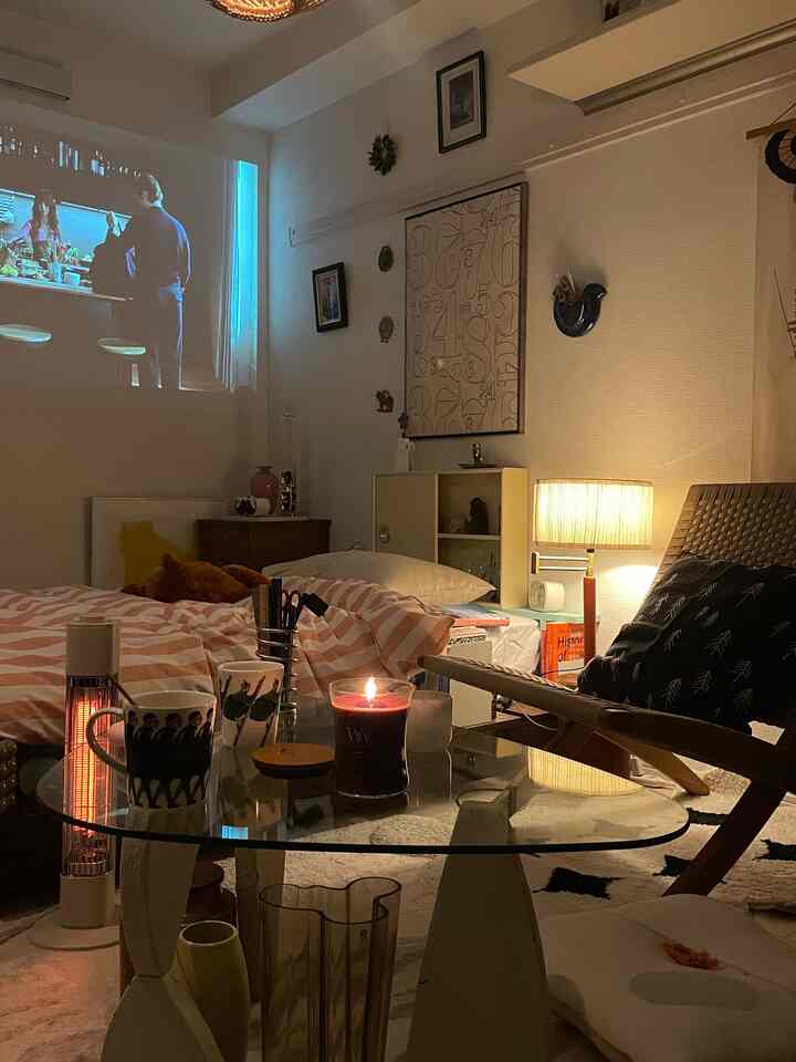 White-toned single household living and bedroom area with warm lighting and pink bedding, glass coffee table featuring a lit candle
