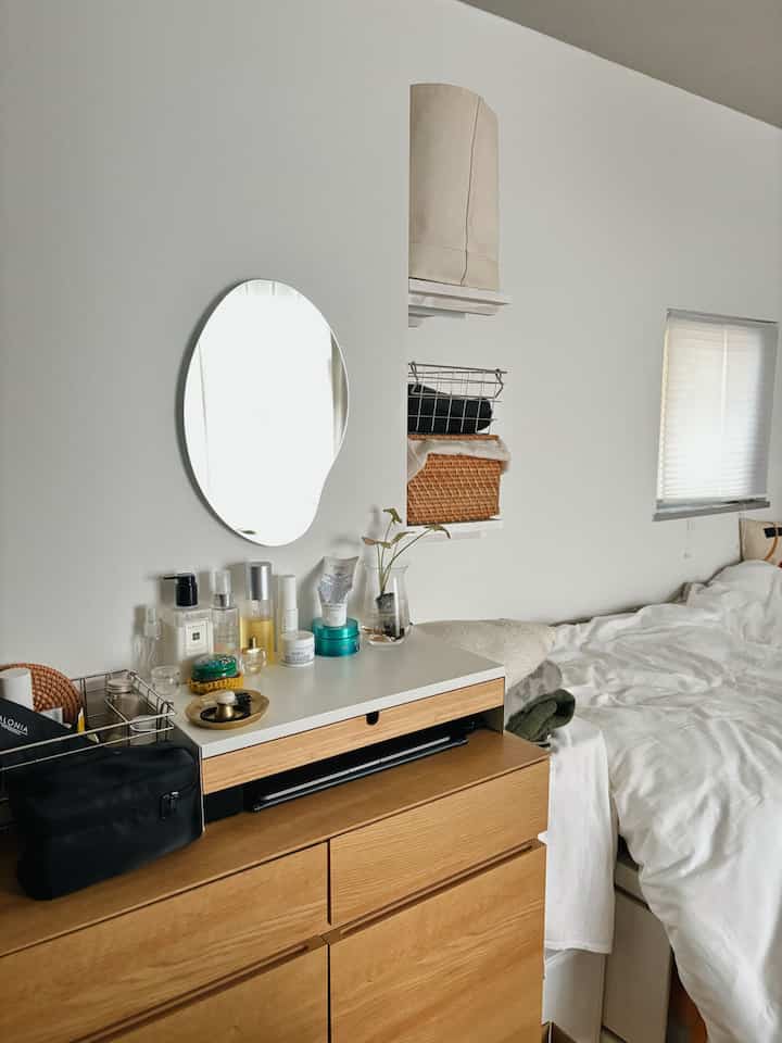 白い壁とナチュラルな木製サイドボードが配置されたワンルームの寝室、壁掛けの丸い鏡と化粧品が整頓された印象的な空間