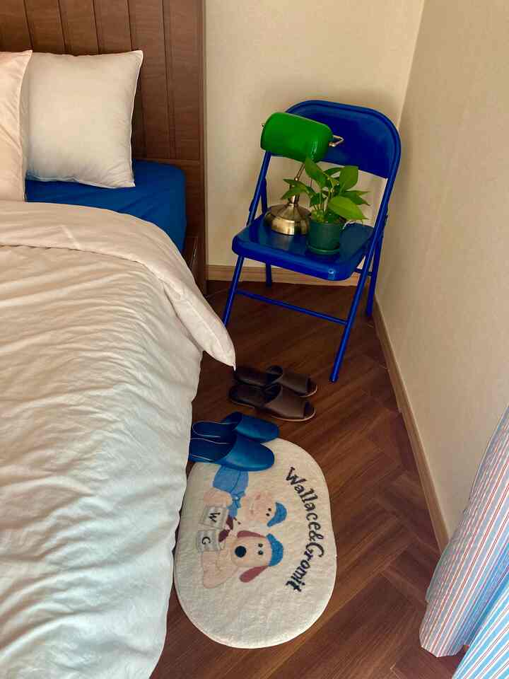 ブラウンの木製フローリングとブルーの寝具がある小さな寝室、青い折りたたみ椅子と白い枕が配置された温かみのある空間