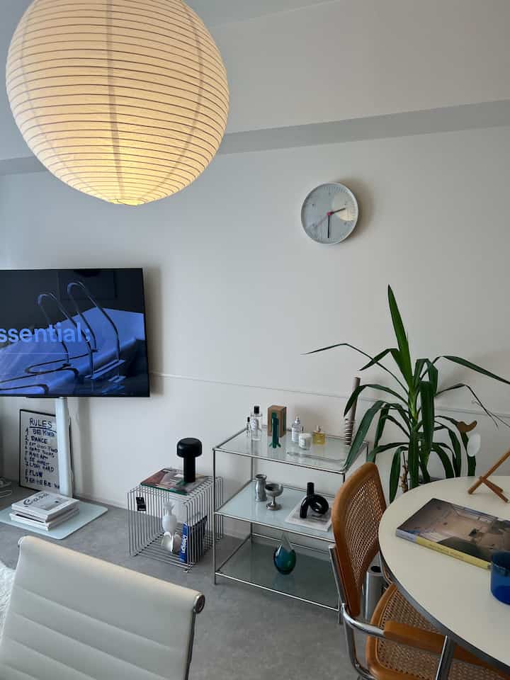 白とナチュラルトーンのリビングルーム、存在感のあるペンダントライトや壁時計、ラタン製のダイニングチェアが配置されたミニマルなインテリア