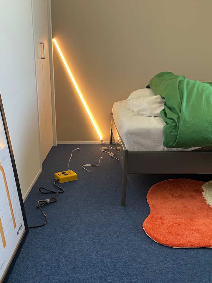 ブルーのタイルカーペットが敷かれたモダンな寝室、グレーのベッドとゴールドのネオン照明が印象的な空間