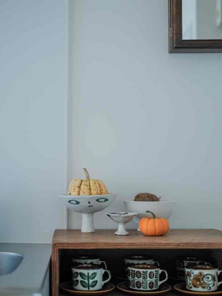 Minimal kitchen space with white walls and wood-tone cabinet, featuring ceramic objets and small pumpkins
