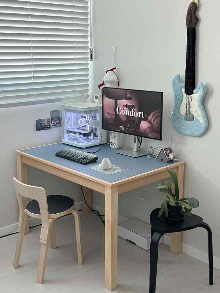 白い壁と木目調の家具が配置されたコンパクトなホームオフィス、机の上のパソコンとスツールの植物があるシンプルなインテリア