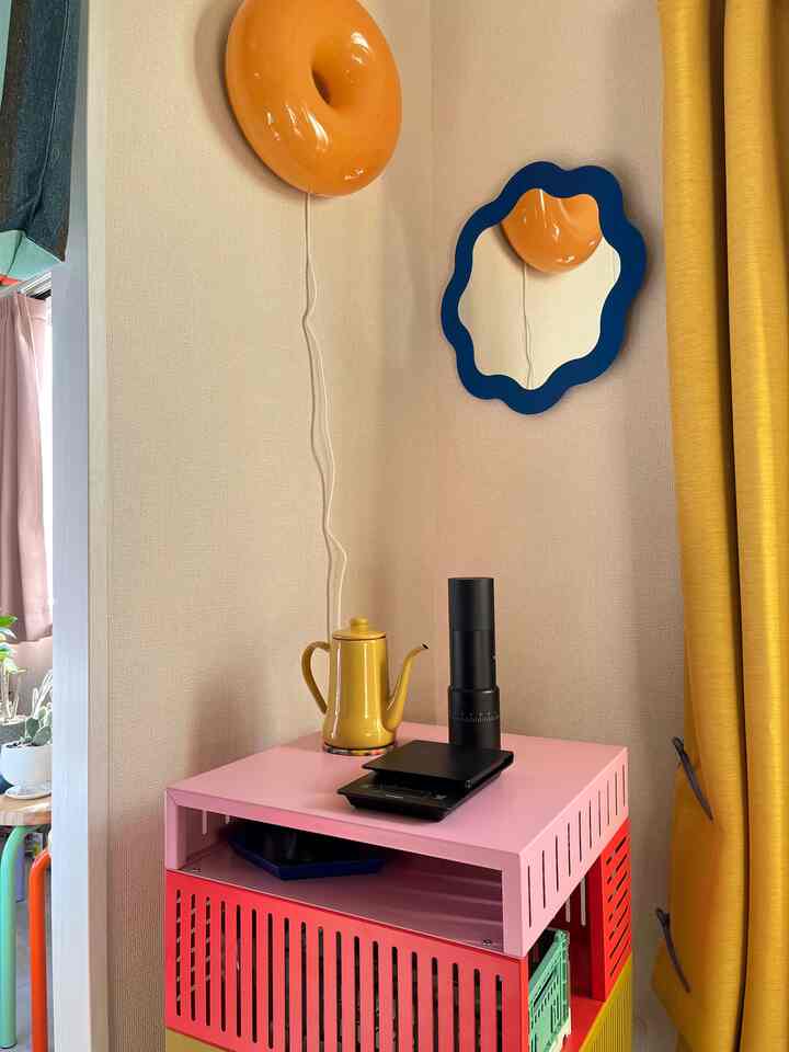 Yellow and pink toned small corner space featuring colorful storage cart, uniquely shaped wall mirror, and exposed wall lighting creating a distinctive home cafe vibe
