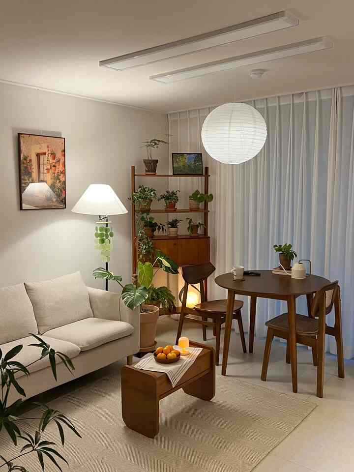 Beige and wood-toned living room featuring a cream sofa, wooden dining set, diverse plants, and soft lighting creating a cozy atmosphere