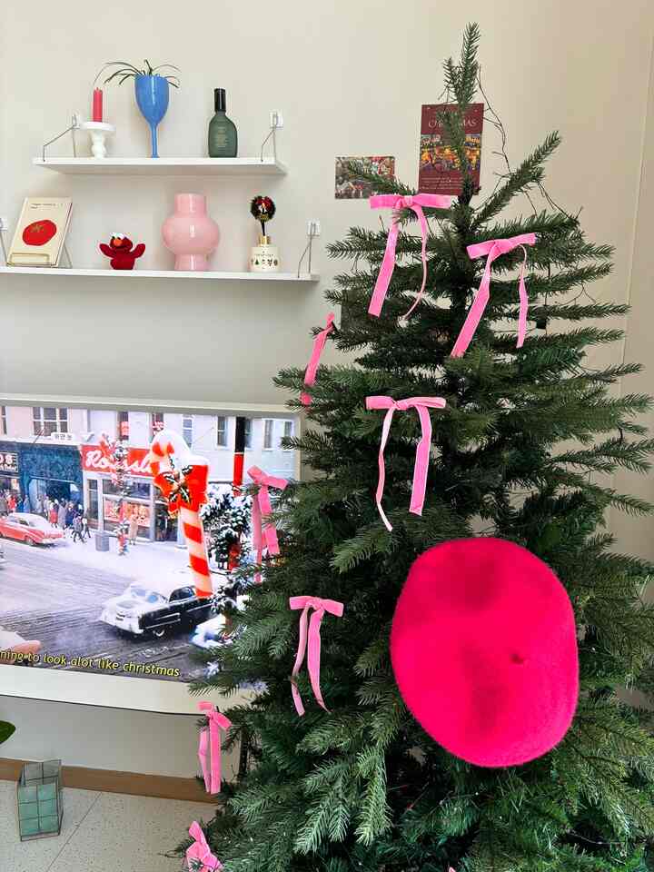 A cozy living room with white walls and vintage furniture, featuring a Christmas tree decorated with pink ribbons and a bright pink beret as the centerpiece