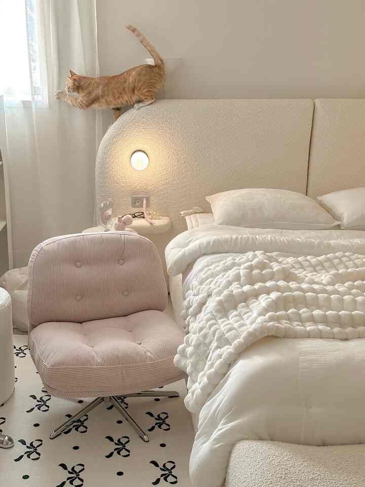 White and natural toned bedroom featuring soft blanket and pink armchair, cozy atmosphere with a cat on the headboard