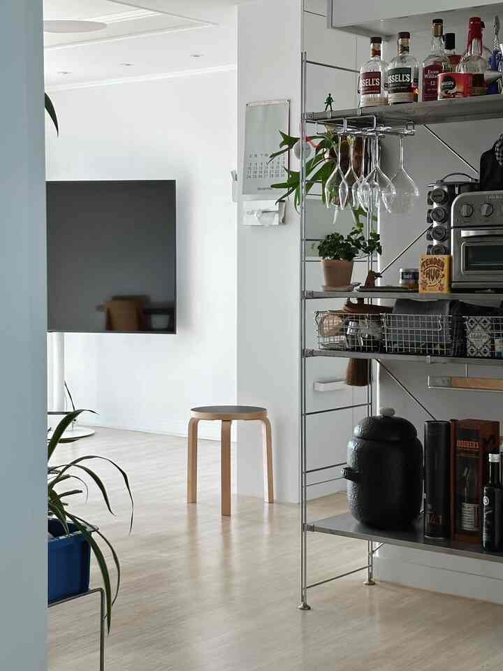 A modern kitchen and living space with white and wood tones, featuring a stool and metal shelves stocked with home bar items, creating a clean atmosphere