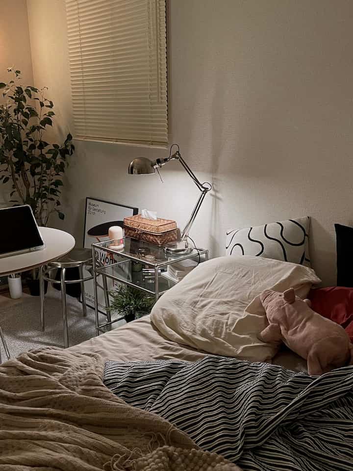 Beige and white toned bedroom featuring bed, transparent nightstand, desk lamp, and plant with a simple natural atmosphere