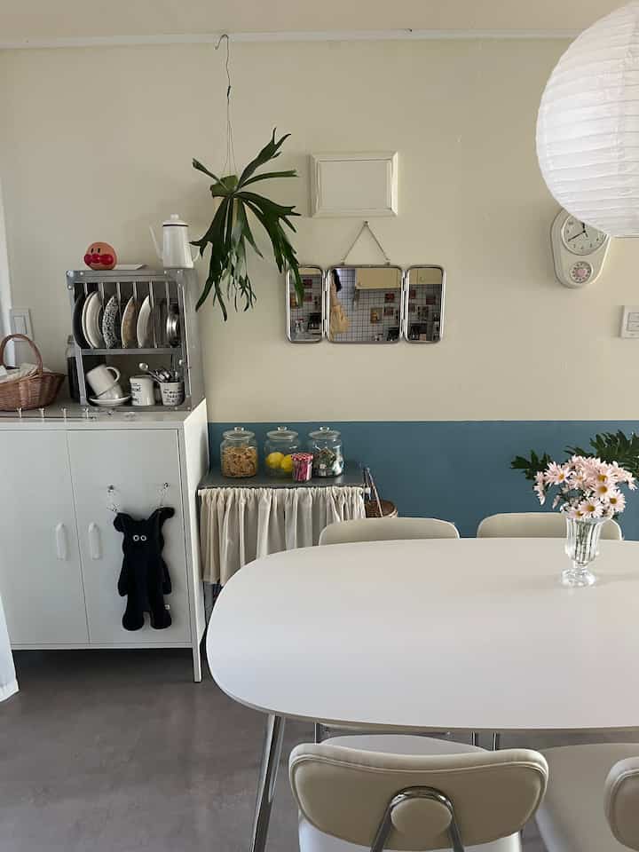 白と青のトーンのキッチンスペース、セラミックのダイニングテーブル、ヴィンテージの三面鏡、コウモリランが配置された雰囲気のあるインテリア