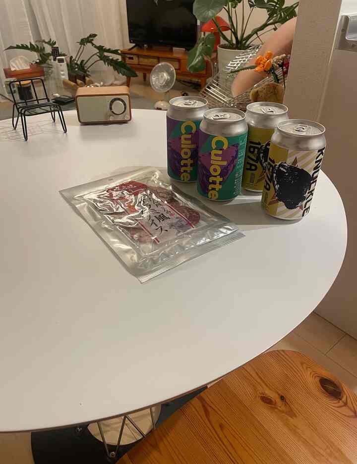 白を基調としたリビング、円形のダイニングテーブルに飲料缶とスナック、ラジオと植物が配置された雰囲気のある空間