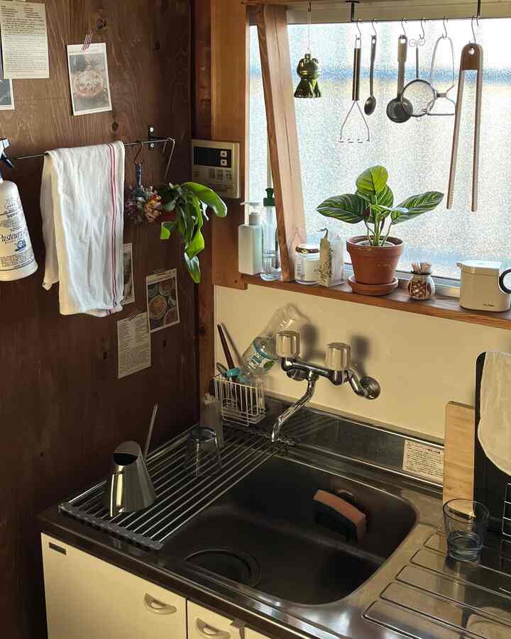 ナチュラルカラーと白を基調としたキッチン、窓際に植物が置かれ、シンクやタオル、調理器具が整った雰囲気のあるインテリア