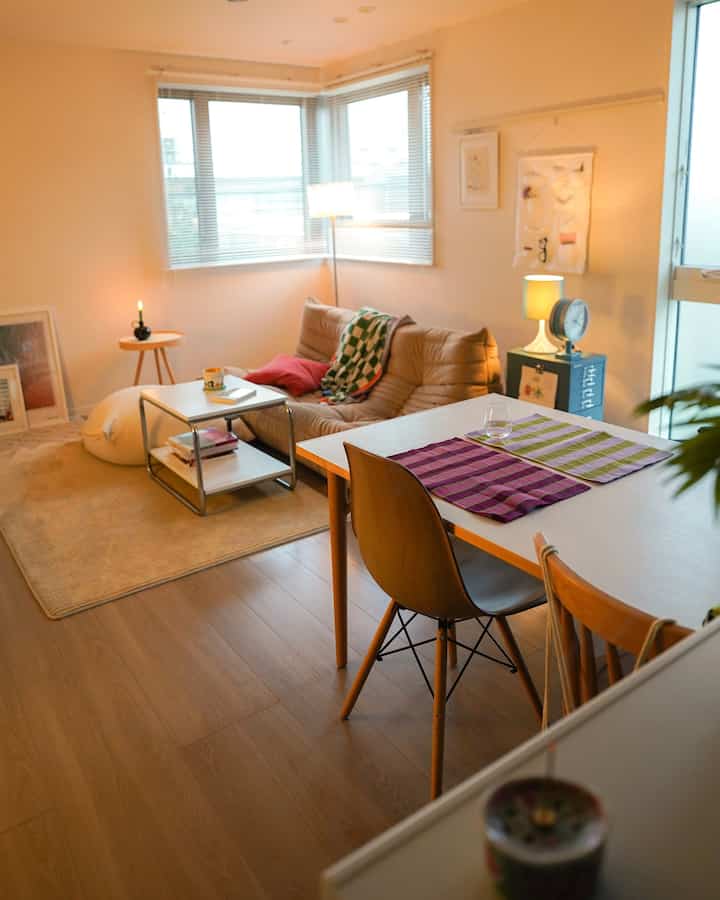 白い壁とナチュラルな木目調の床があるワンルームのリビングとダイニングスペース、ベージュのソファとシンプルな家具が配置された暖かみのあるインテリア