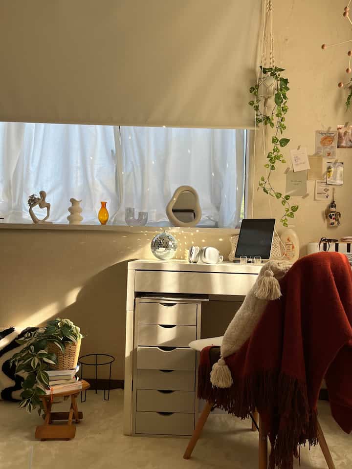 明るいナチュラルカラーの小さなホームオフィス。白いデスクと赤いブランケットのかかった椅子、植物が配置された温かみのあるインテリア