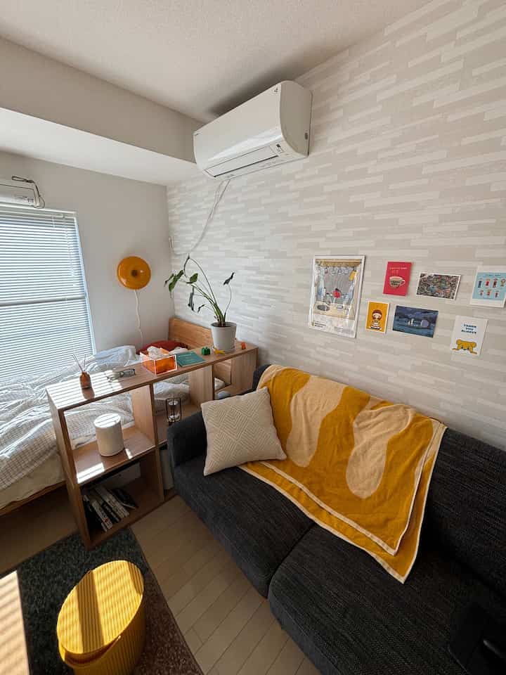 ベージュの壁と黄色のアクセントがある7畳のワンルームリビング、ソファとベッド、木製収納棚が配置された落ち着いた空間