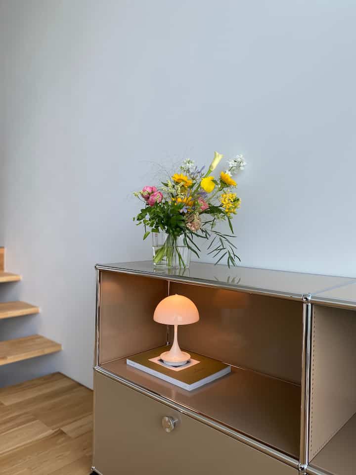 明るい白い壁と茶色い木の床のモダンな玄関に、サイドボード、テーブルランプ、生花の花瓶が配置されたナチュラルな空間