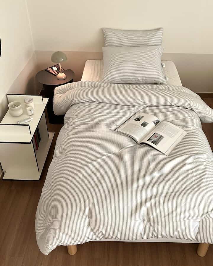 グレーと白のモノトーン寝室、ストライプ柄のマイクロファイバー寝具とテーブルランプが配置されたシンプルモダンな印象的な空間