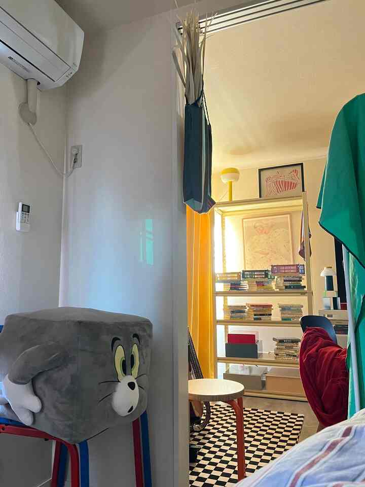 黄色のカーテンと黒のチェック柄ラグがあるコンパクトで狭い部屋、本棚とスツールが配置された温かみのあるインテリア