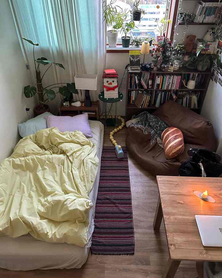 ウッドトーンの床と黄色の寝具がある小さな寝室、本棚とビーンバッグ、植物が配置された居心地の良い空間