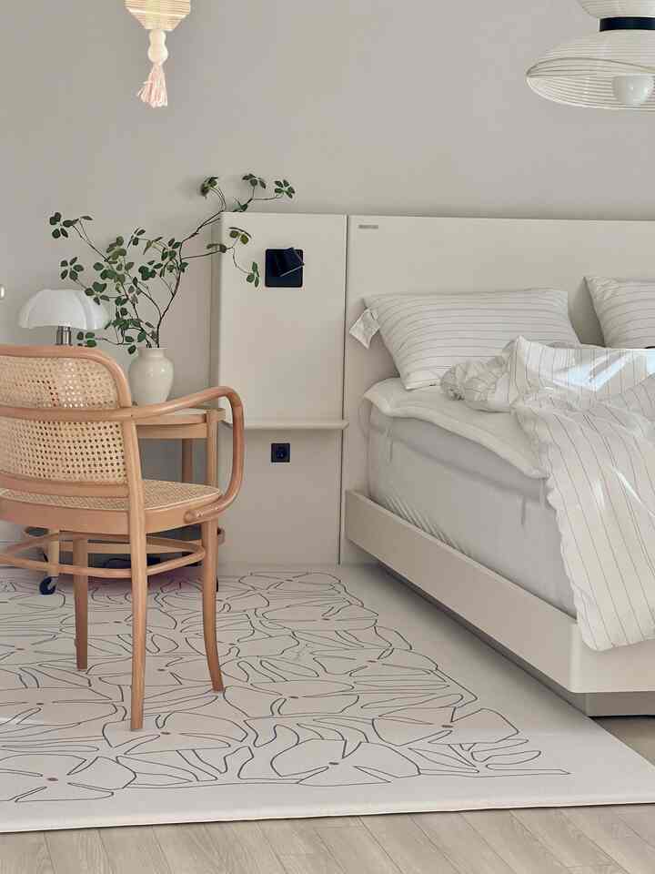 White-toned bedroom featuring a white bed with pillows, natural wood armchair, leaf-patterned rug, and a plant in a vase, creating a clean and natural atmosphere